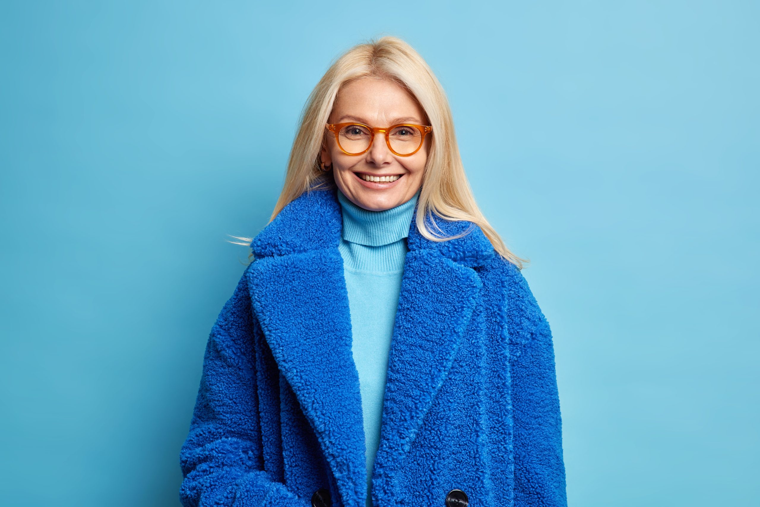 Smiling European woman with blonde hair dressed in blue winter coat has happy mood wears eyewear poses in studio. Fashionable middle aged blonde lady in outerwear going to have walk with family